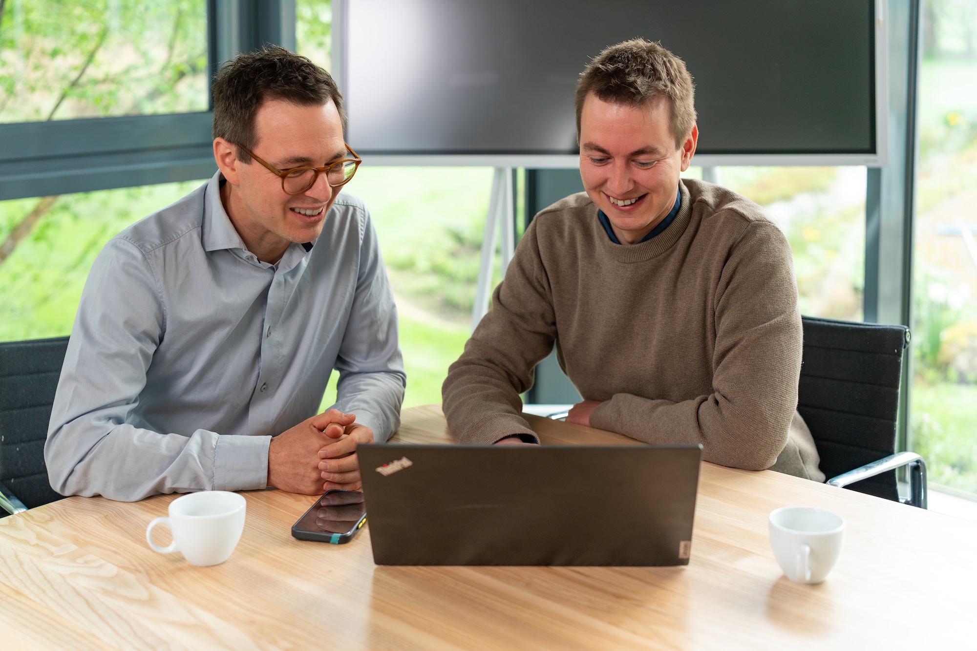 Zwei Männer sitzen an einem Tisch und schauen lächelnd auf einen Laptop.