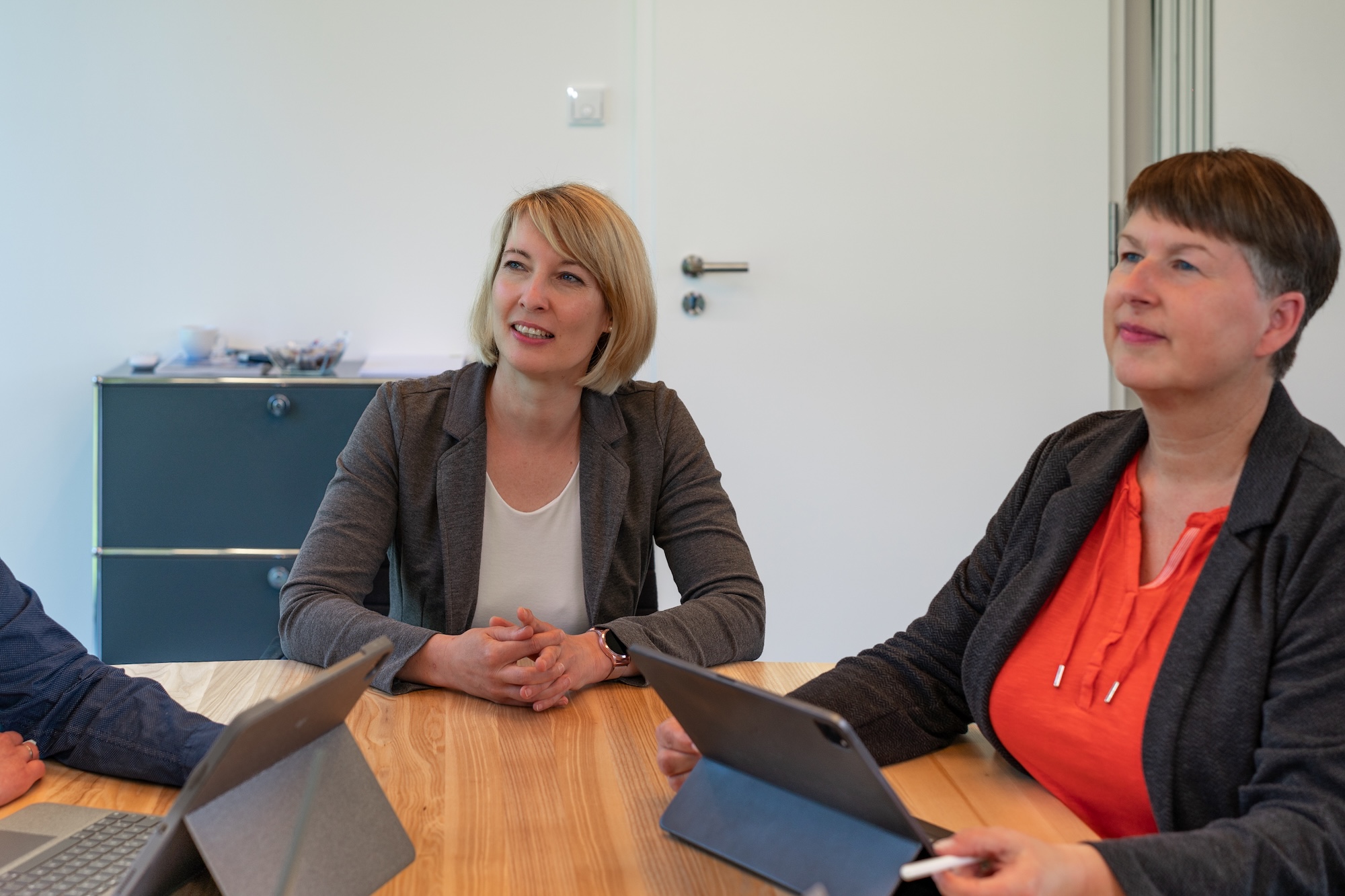 Zwei Frauen sitzen an einem Holztisch mit Tablets und blicken aufmerksam in eine Richtung.