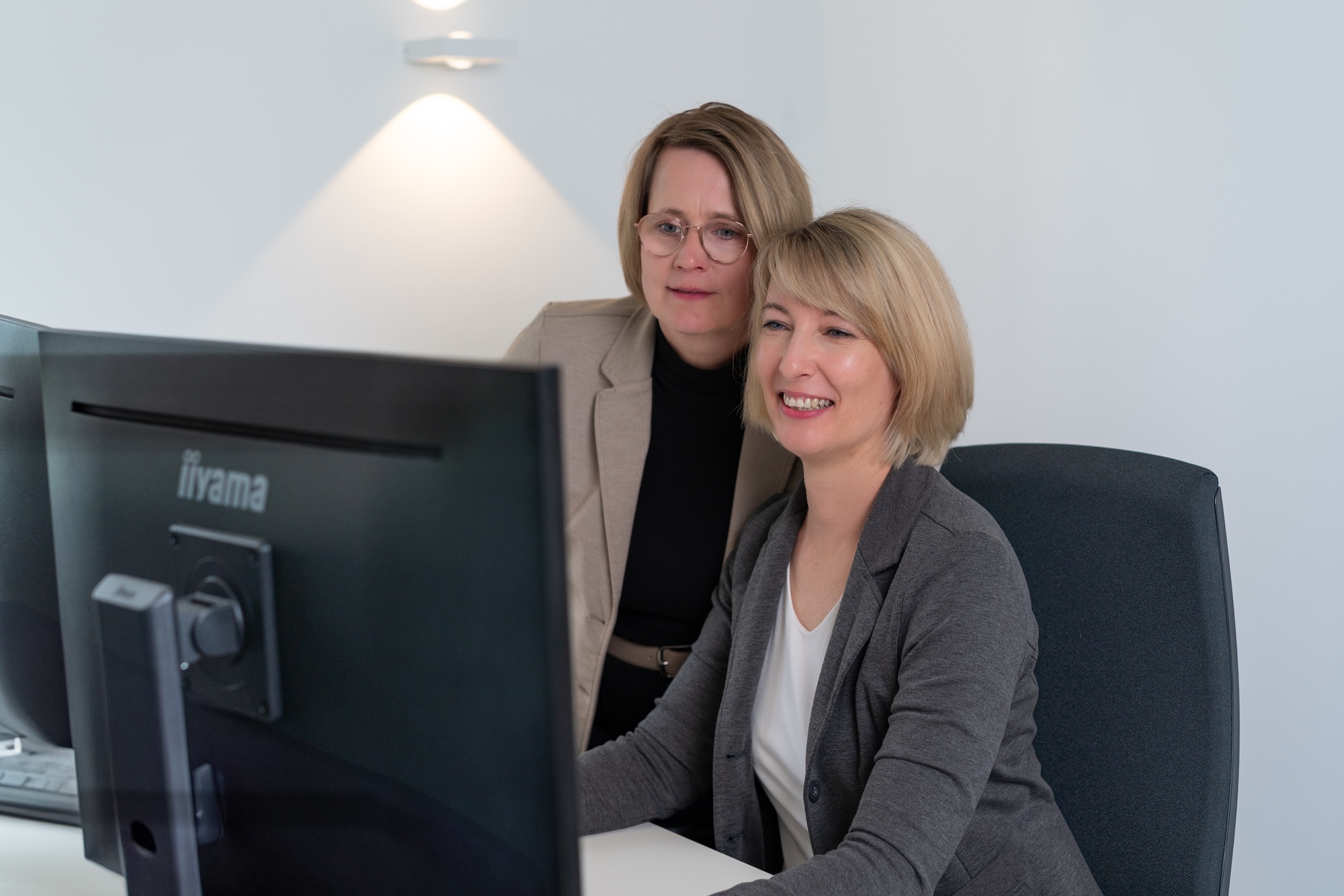 Zwei Frauen lächeln und schauen gemeinsam auf einen Computerbildschirm in einem hellen Büro.