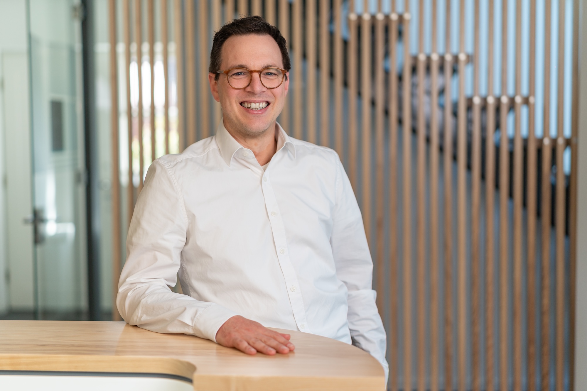 Mann mit Brille und weißem Hemd lächelt stehend an einem hellen Holztisch, Hintergrund mit vertikalen Holzlatten.