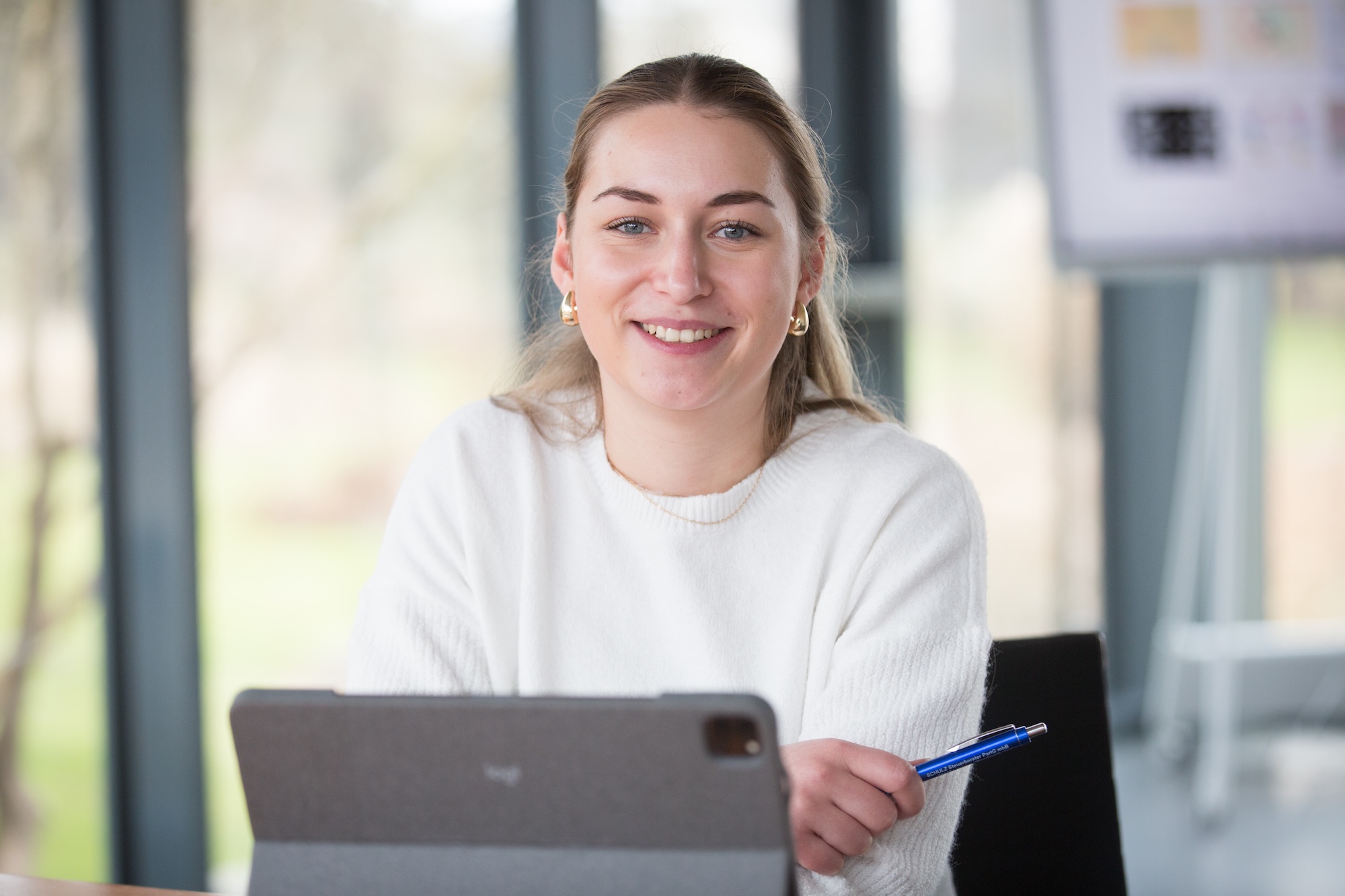 Junge Frau in weißem Pullover, die lächelt und einen Stift hält, während sie vor einem Tablet sitzt.