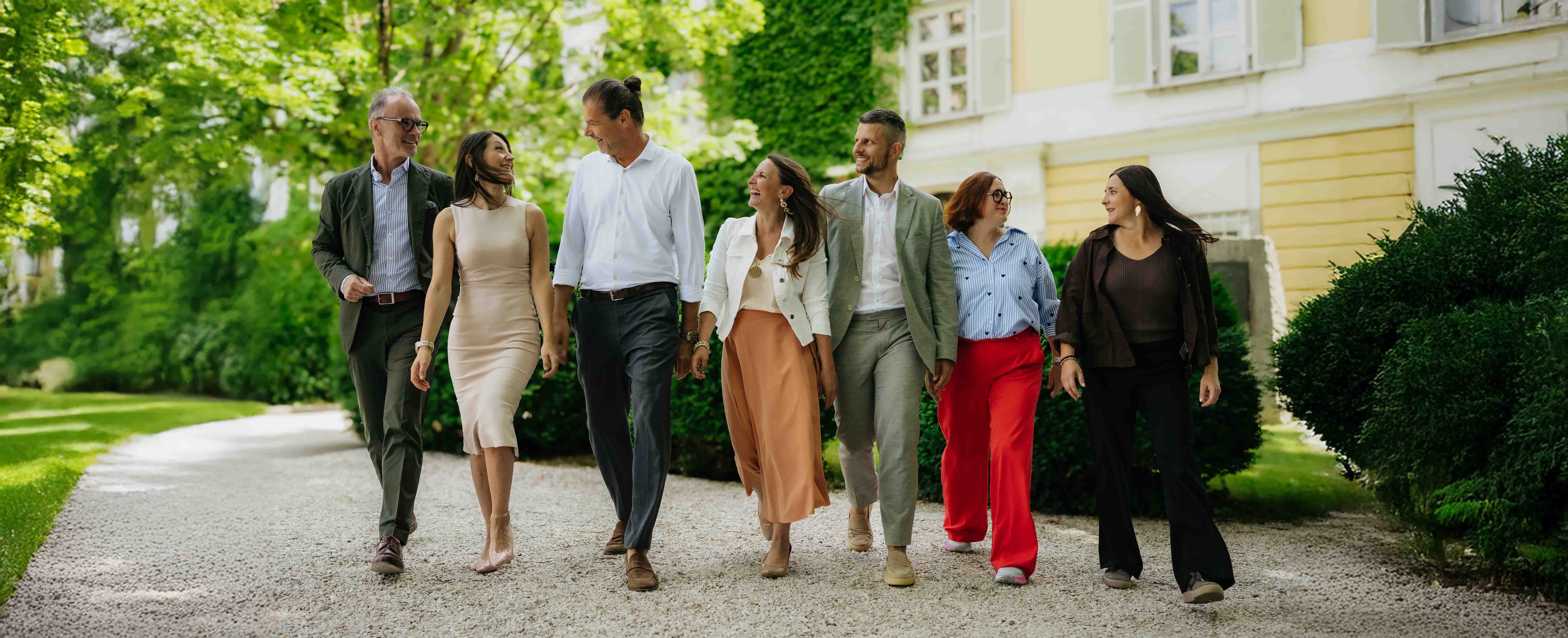 Sechs Personen gehen lachend auf einem Kiesweg mit Gebäuden und Bäumen im Hintergrund.