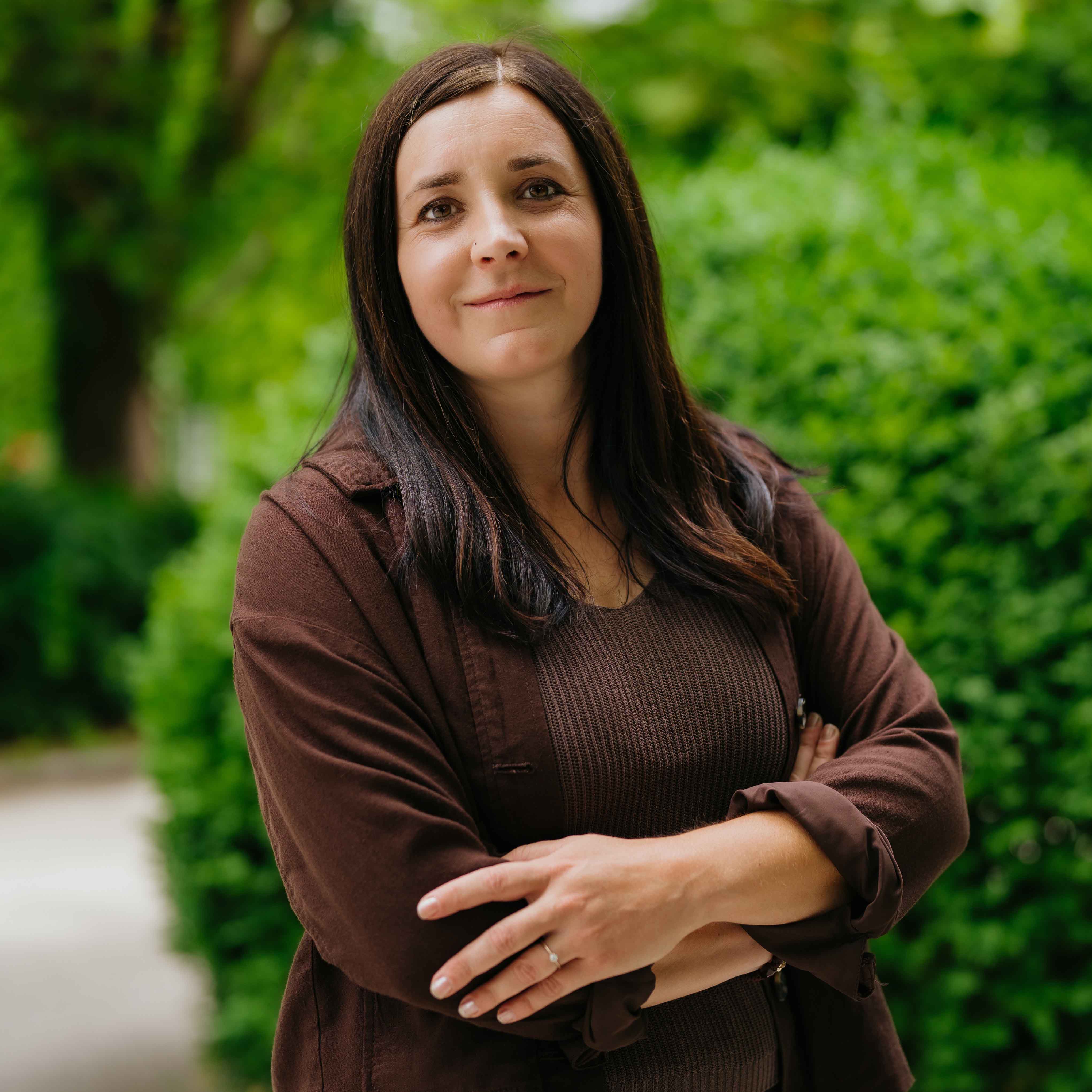 Frau mit langen braunen Haaren und brauner Kleidung steht vor grünen Büschen im Freien.