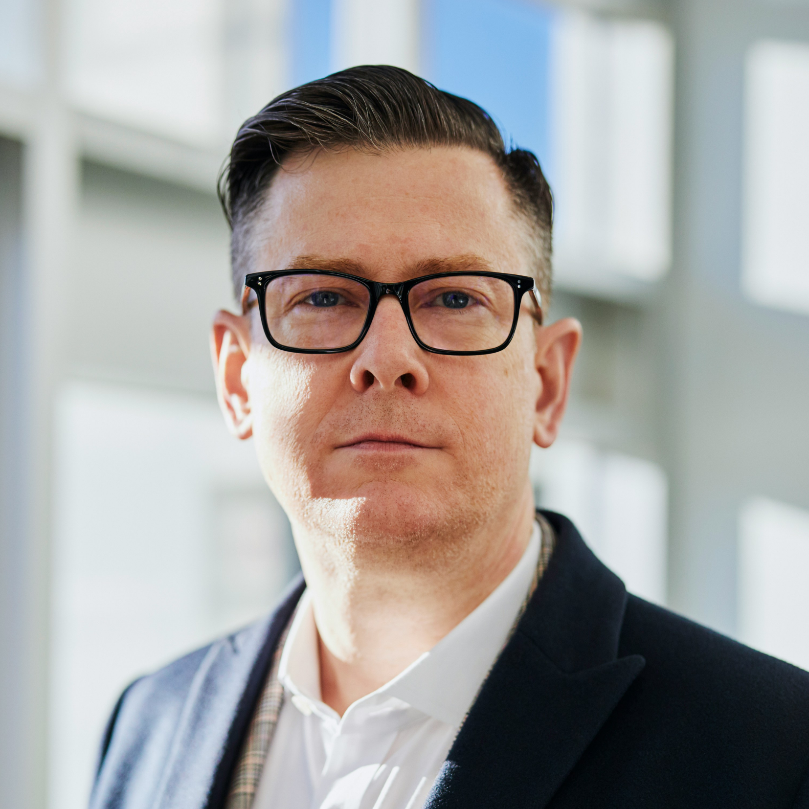 Man with black glasses wearing a dark blazer and white shirt in a bright indoor setting.