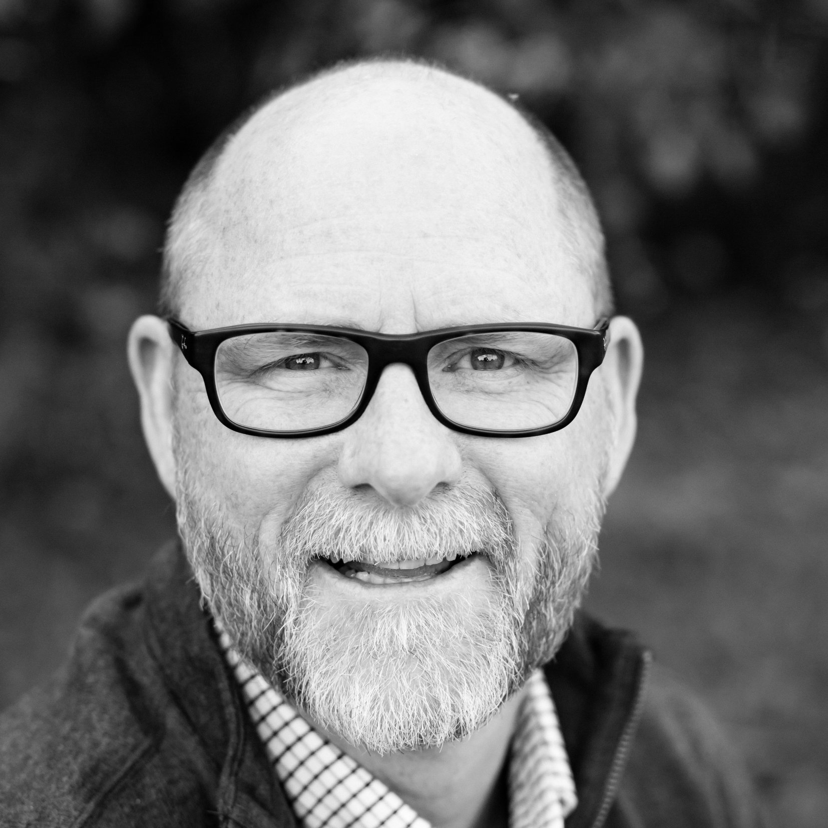 Smiling bald man with glasses, beard, and checkered shirt in a black and white close-up portrait.
