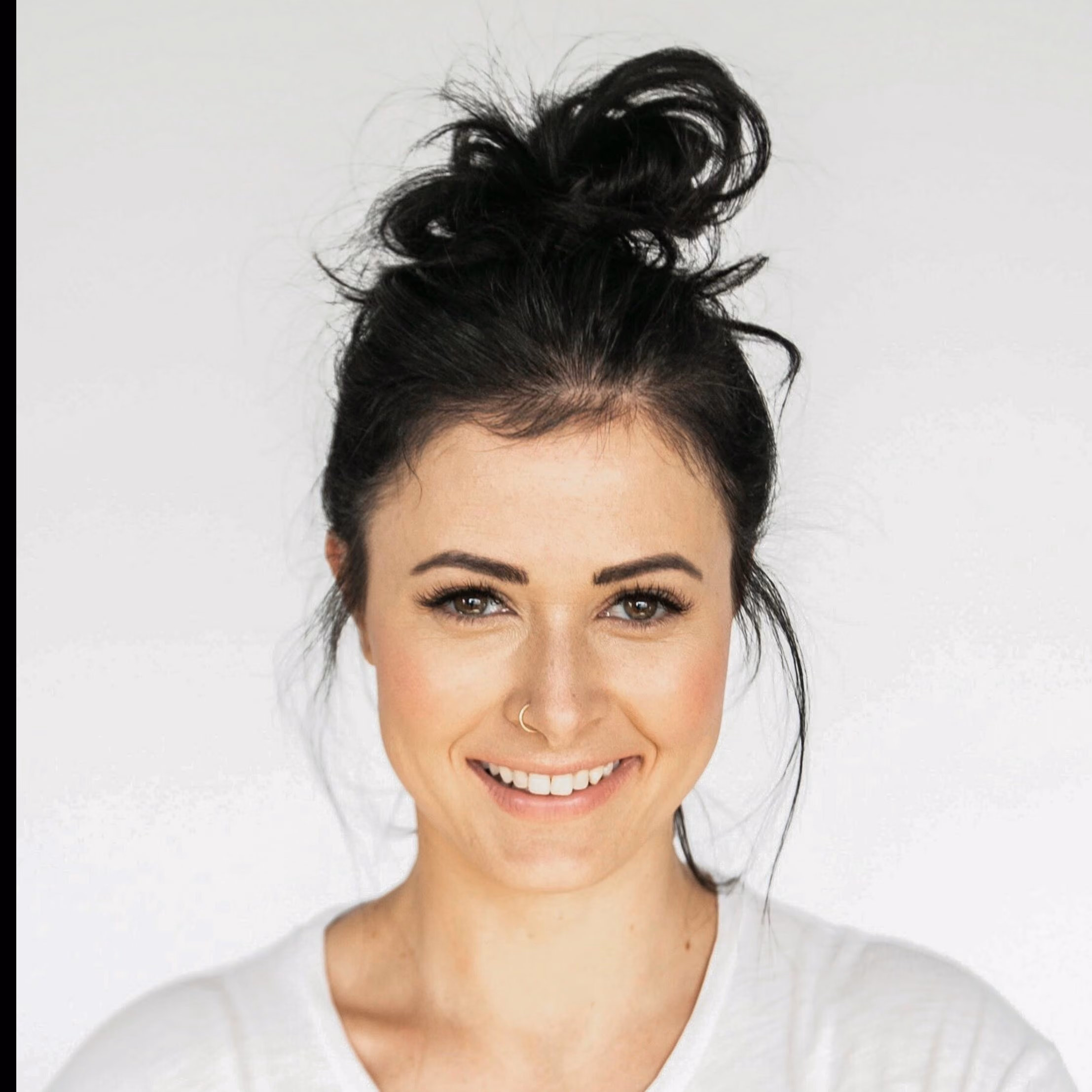 Smiling woman with dark hair in a messy bun wearing a white top and a nose ring.