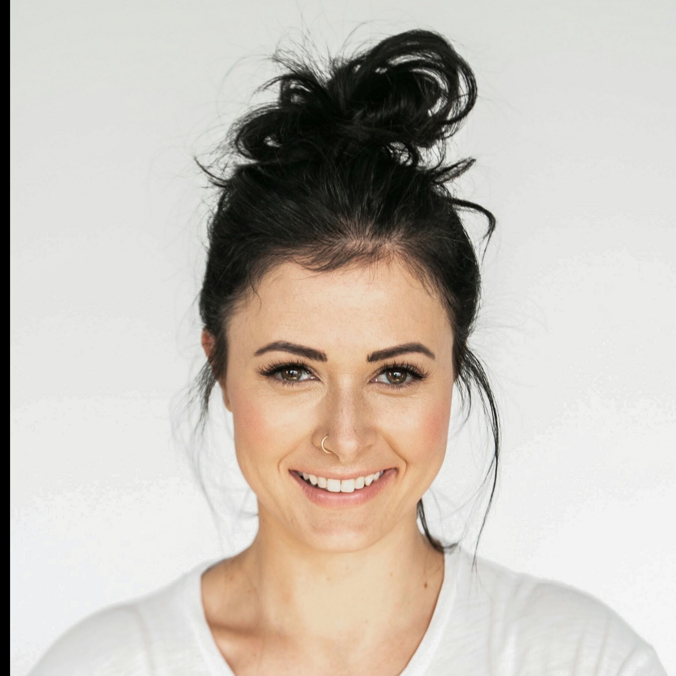 Smiling woman with dark hair in a messy bun wearing a white top and a nose ring.