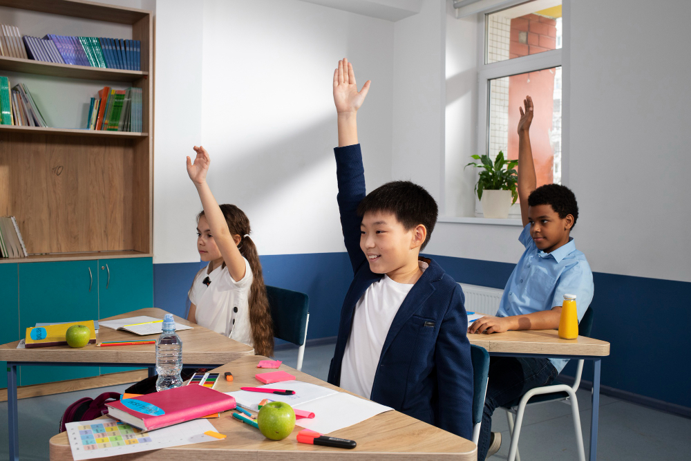 Three schoolchildren sitting at desks in classroom, smiling with raised hands to answer a question.