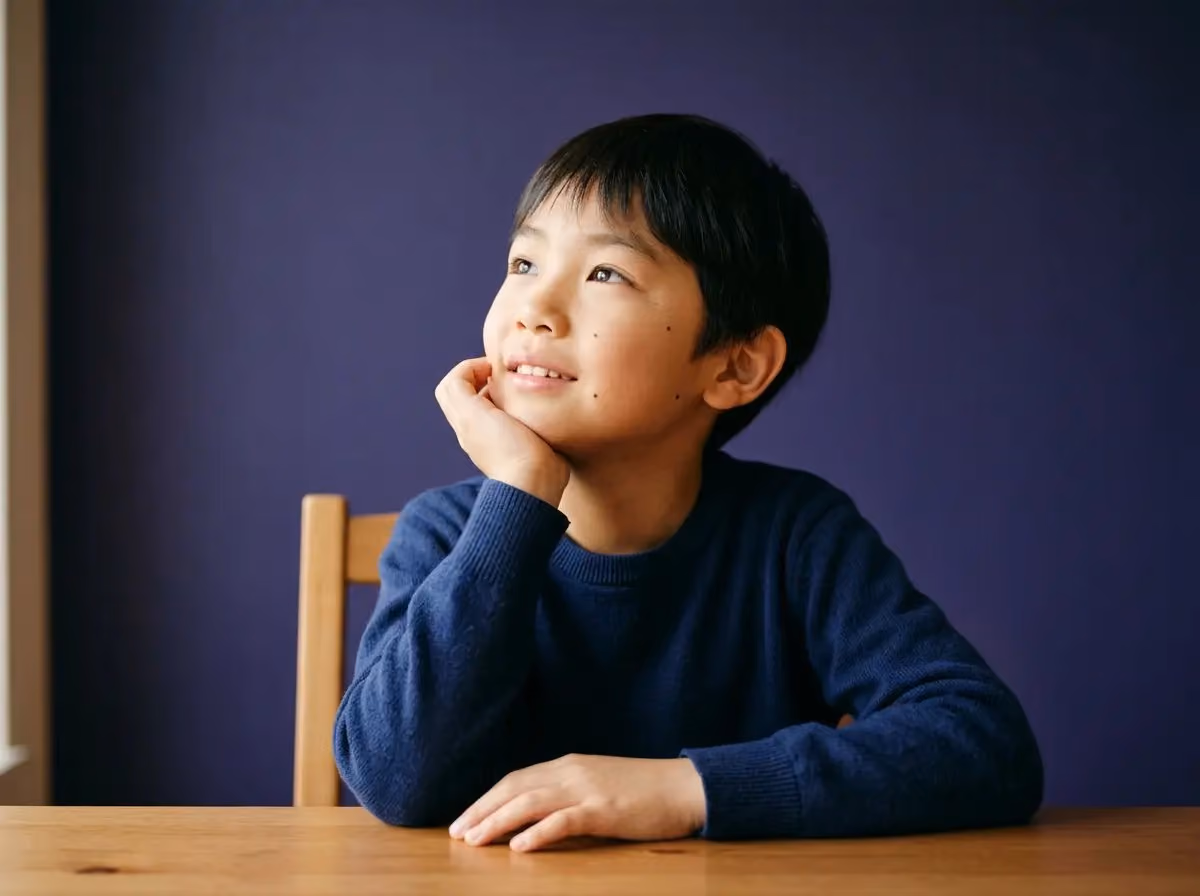 Young boy in navy sweater resting chin on hand and looking up, seated at wooden table with purple wall.