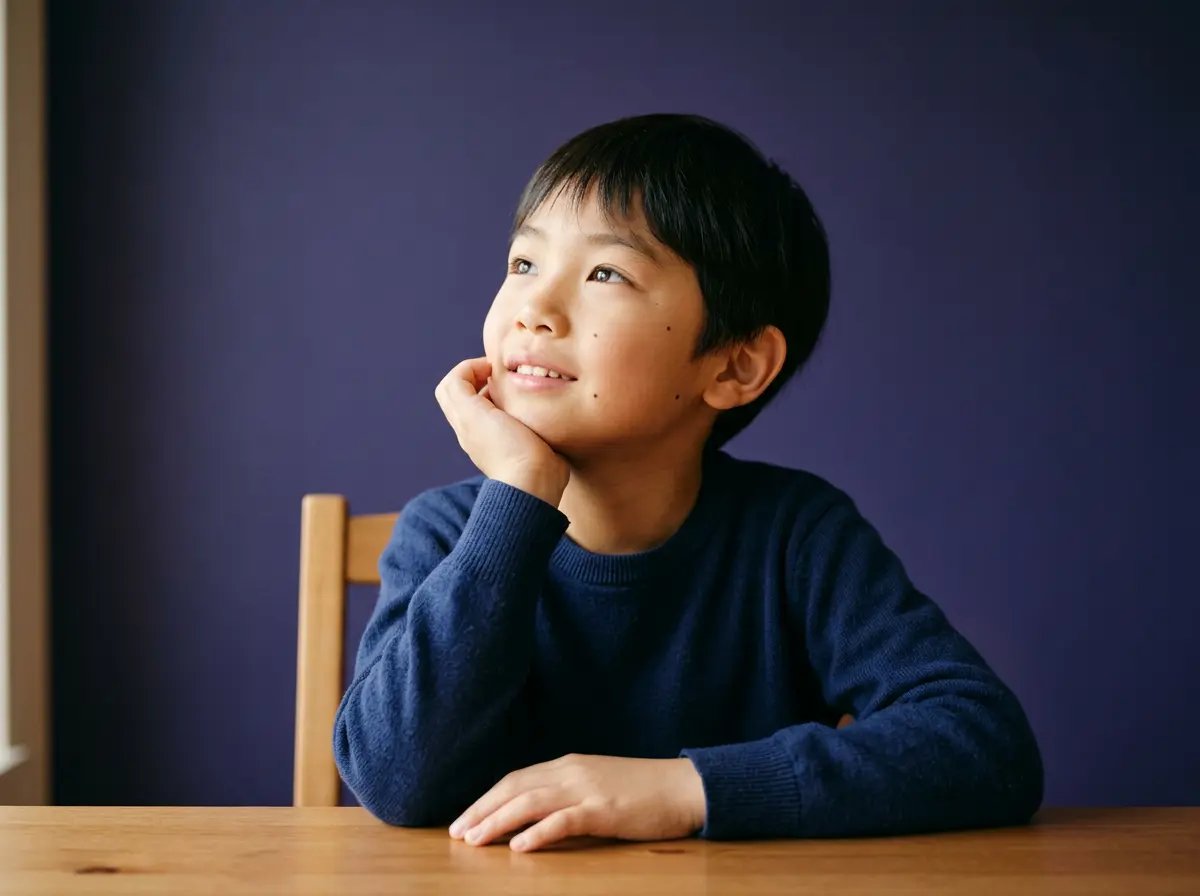 Young boy in navy sweater resting chin on hand and looking up, seated at wooden table with purple wall.