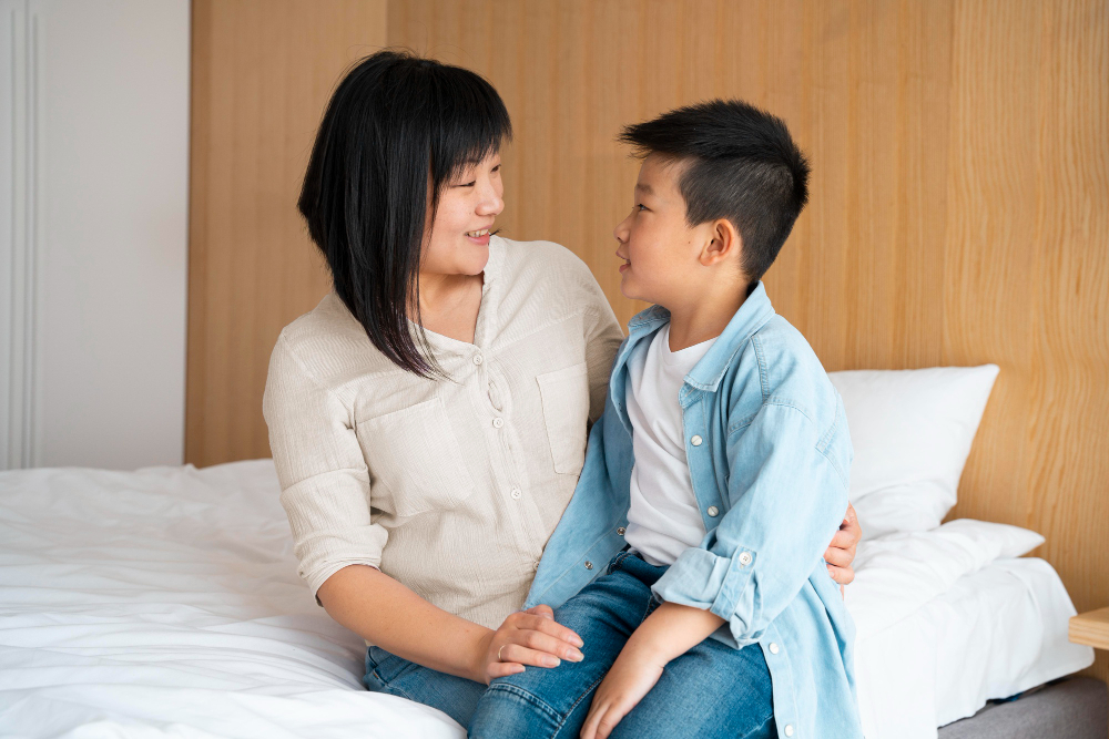 Mother and son sitting on bed looking at each other and smiling in a bedroom with wooden headboard.