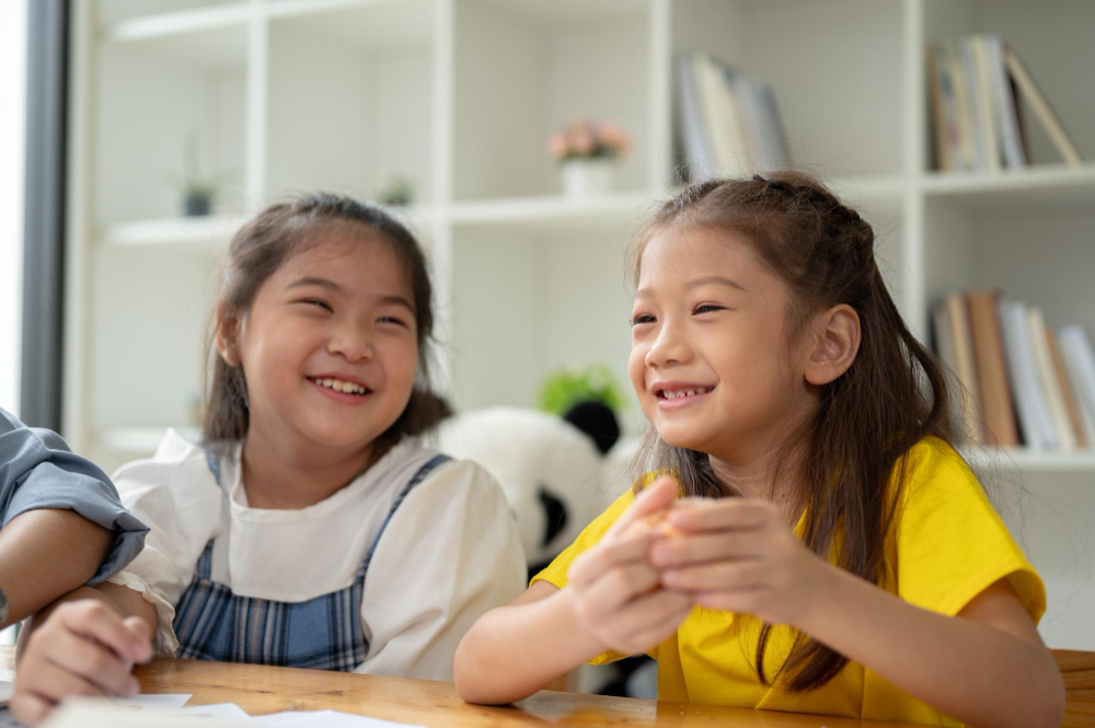 Two young girls smiling and enjoying time together indoors at a table with shelves in the background.