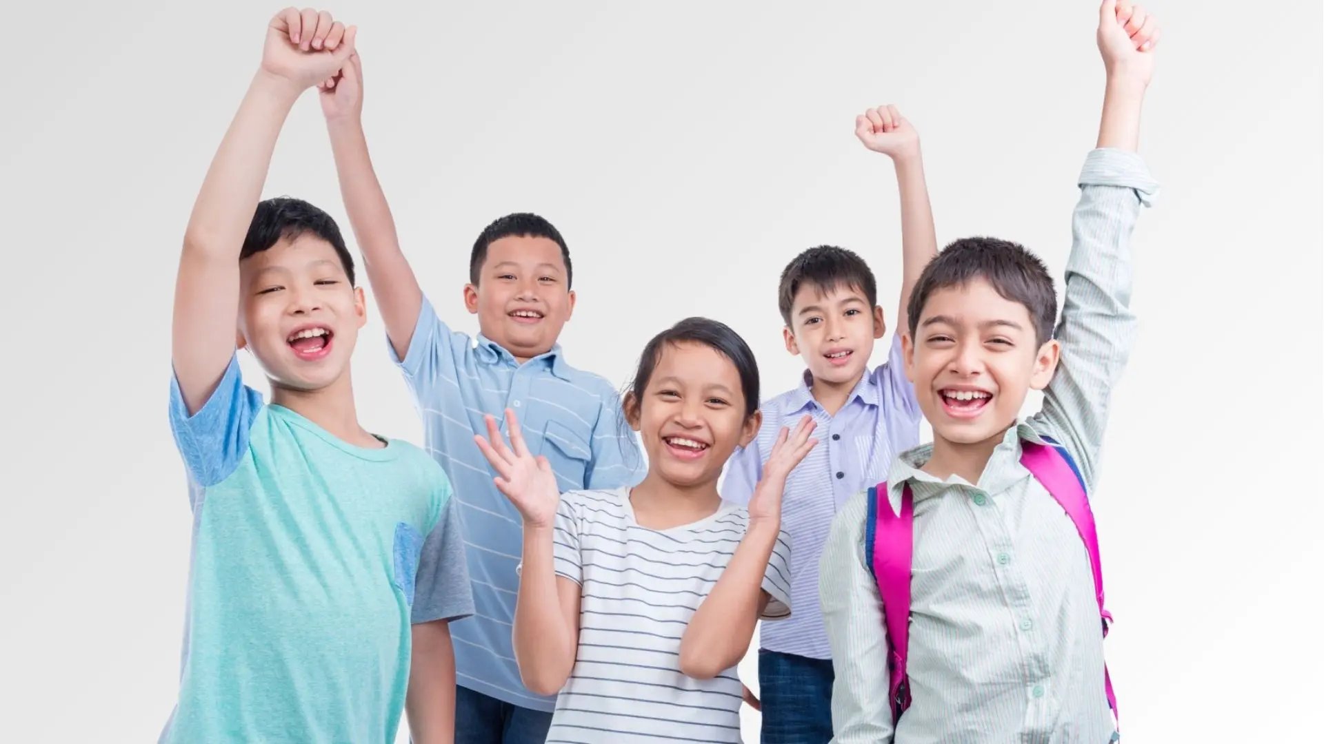 Five happy children smiling with some raising their hands, standing against a white background.