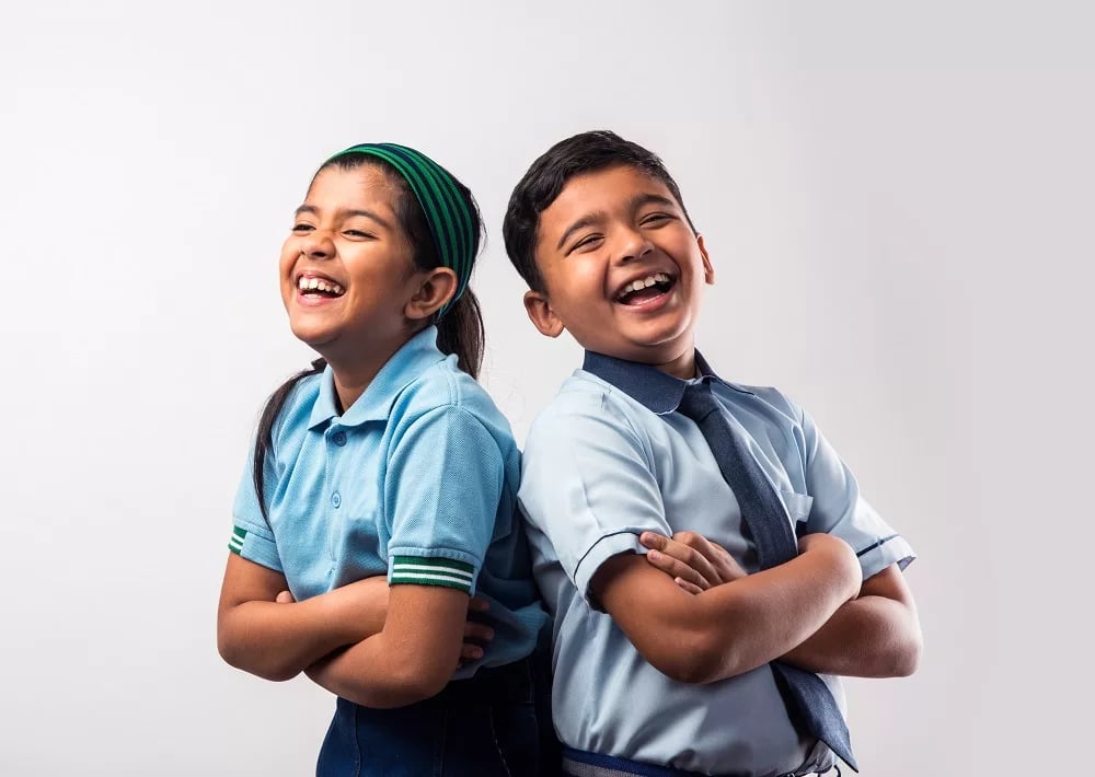 Two schoolchildren in uniforms laughing with arms crossed, standing back to back against a white background.