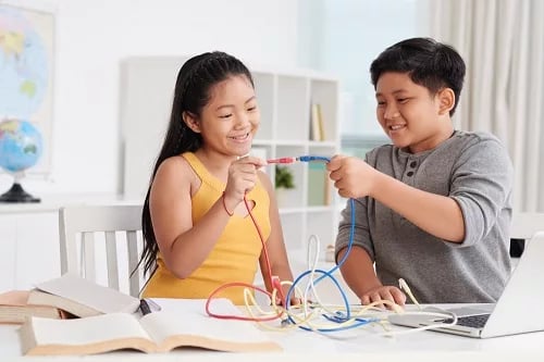Two children smiling and holding wires while working on a science project at a desk with books and laptop.