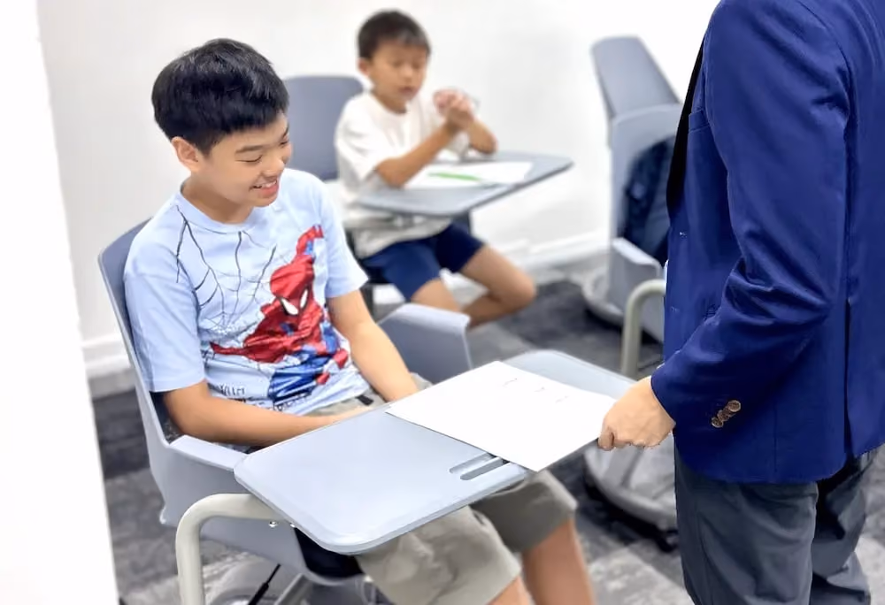 Student in a Spider-Man shirt receives a paper from a standing adult in a blue blazer in a classroom.