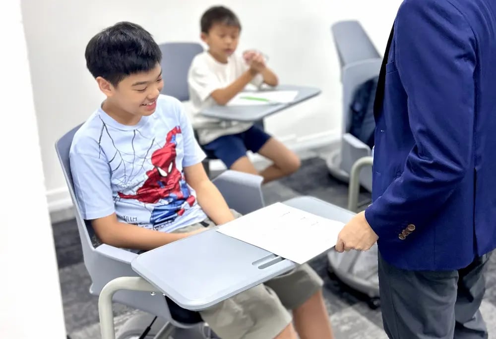 Student in a Spider-Man shirt receives a paper from a standing adult in a blue blazer in a classroom.