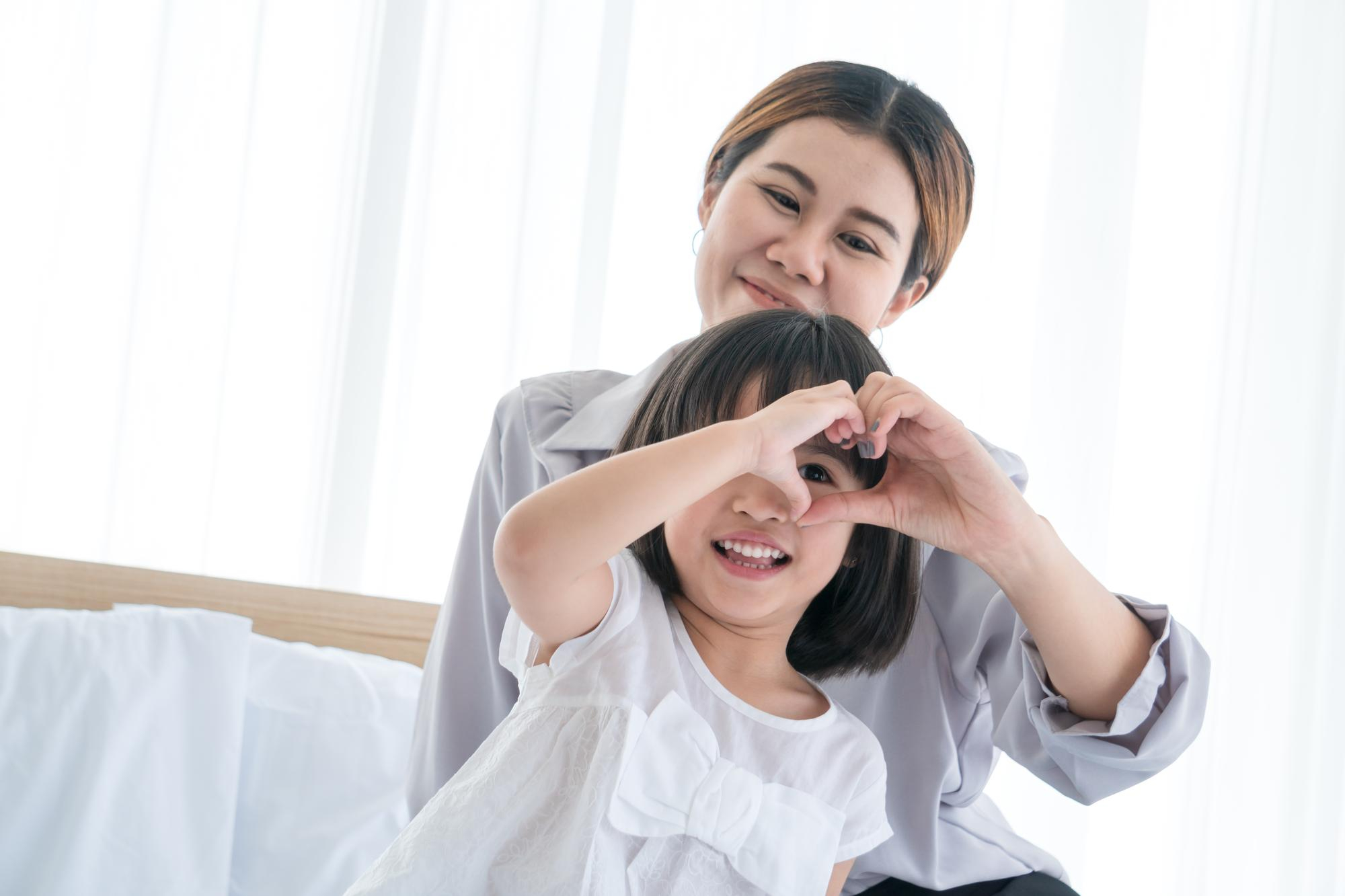Smiling child making a heart shape with hands in front of a woman in a light room.
