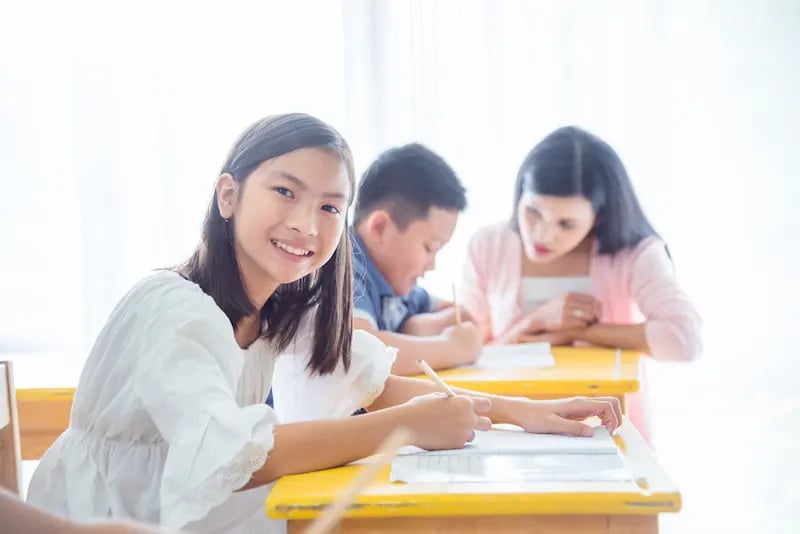 Young girl smiling at camera while writing at a desk, with a boy and woman working behind her.