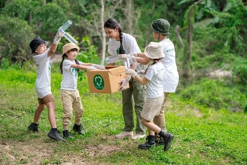 Woman and four children outdoors holding a recycling box and empty plastic bottles in a green park.