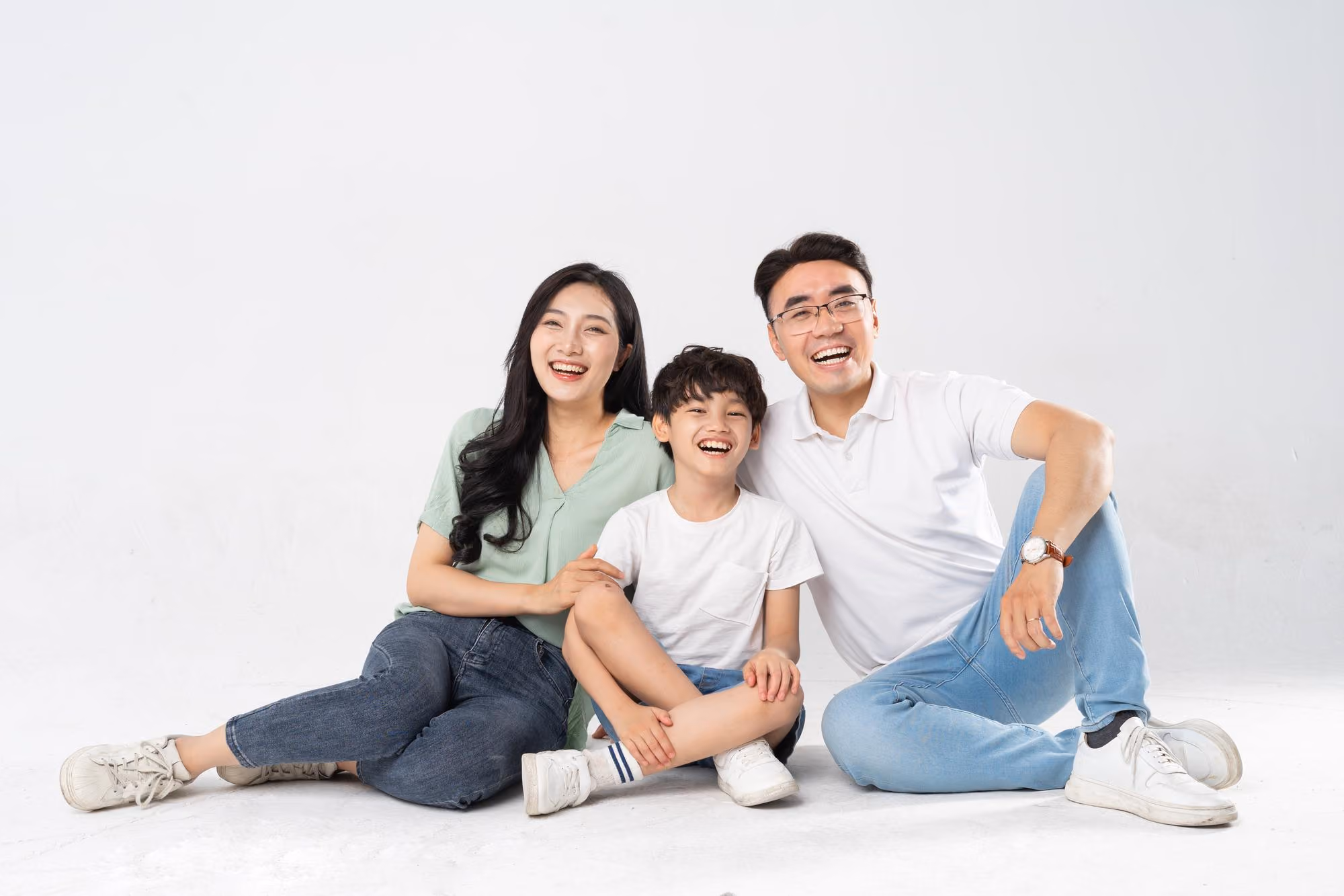 Happy Asian family of three sitting on floor together, smiling and laughing against white background.