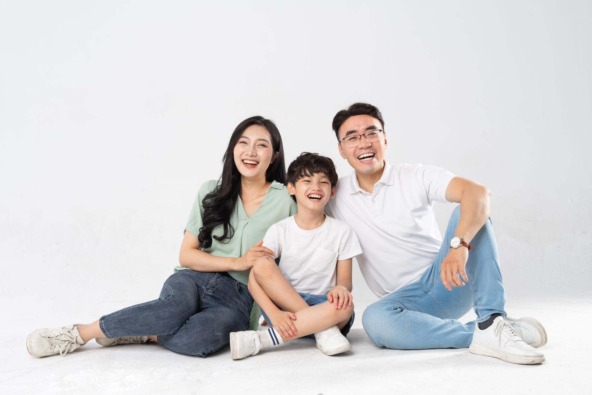 Happy Asian family of three sitting on floor together, smiling and laughing against white background.