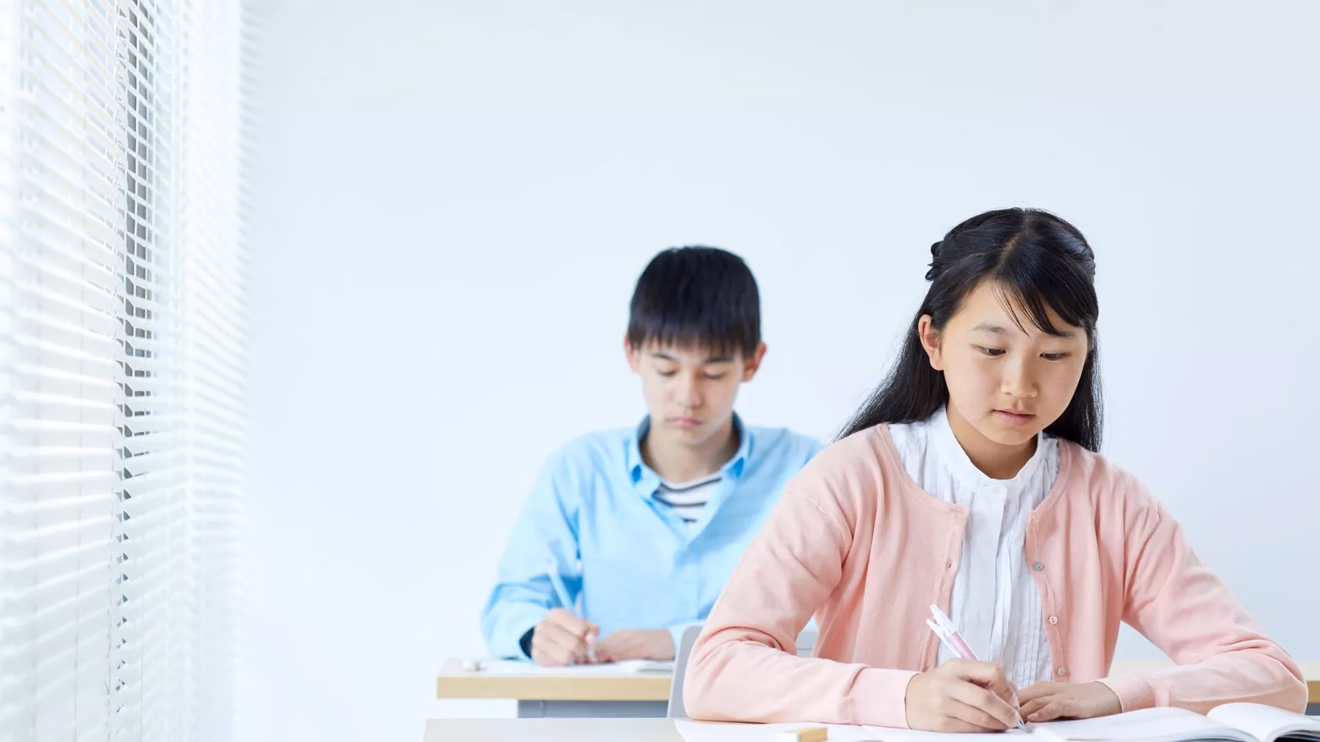 Two students sitting at desks writing in notebooks in a bright classroom by a window.