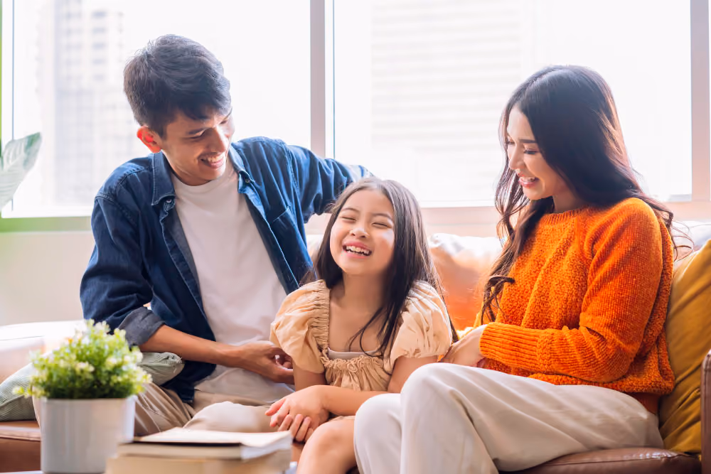 Smiling Asian family of three sitting on couch, enjoying time together in bright living room.
