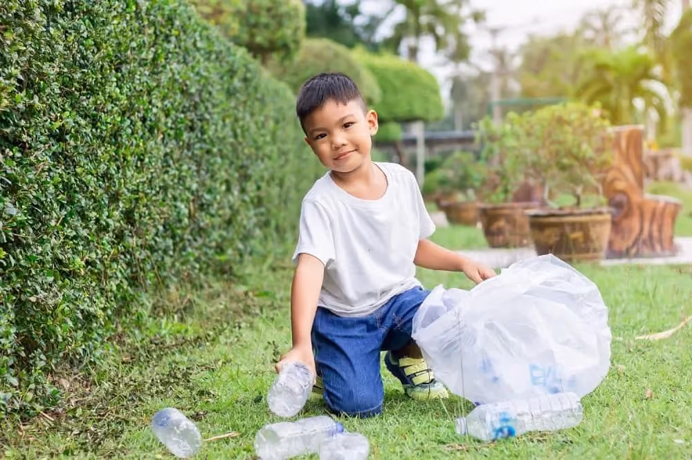 Young boy kneeling on grass, collecting plastic bottles in a large transparent bag outdoors.