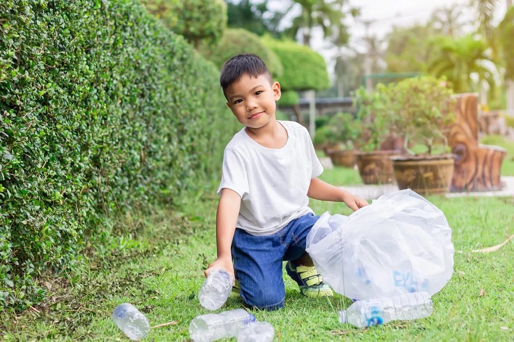 Young boy kneeling on grass, collecting plastic bottles in a large transparent bag outdoors.