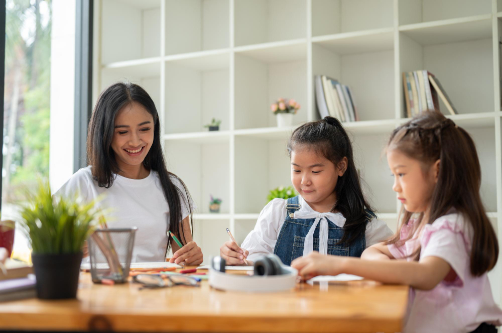 Young woman and two girls drawing together at a wooden table in a bright room with shelves.