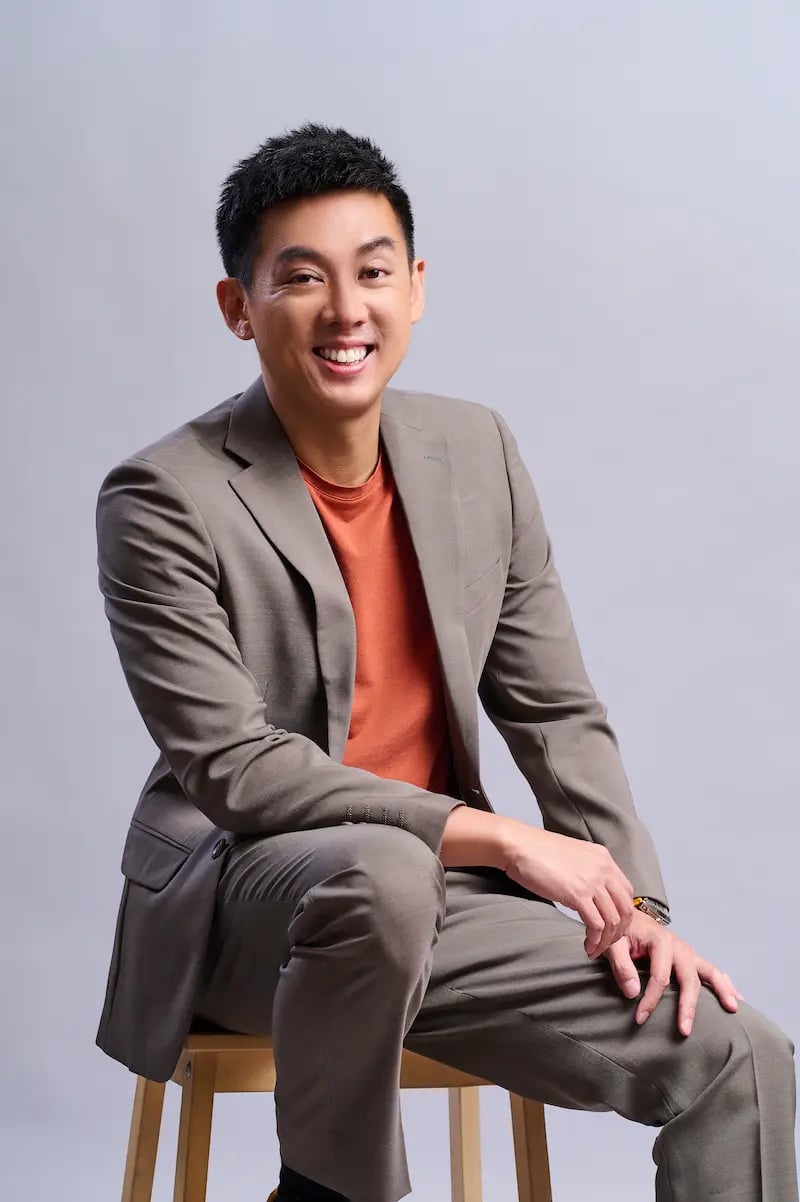 Smiling man in a brown suit and orange shirt sitting on a wooden stool against a gray background.