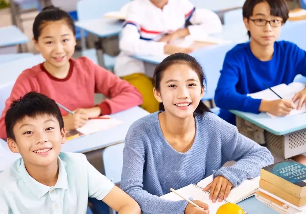 Smiling students sitting at desks with notebooks and pencils in a classroom setting.