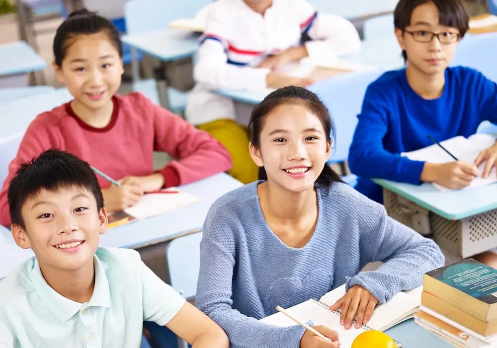 Smiling students sitting at desks with notebooks and pencils in a classroom setting.