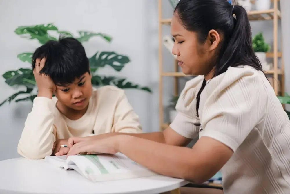 Two young Asian children sitting together, focused on a textbook while studying at a table.