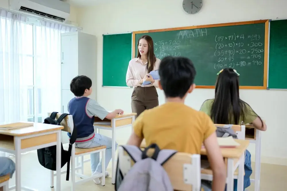 Asian female teacher standing at a green chalkboard while children sit at their desks in a classroom.