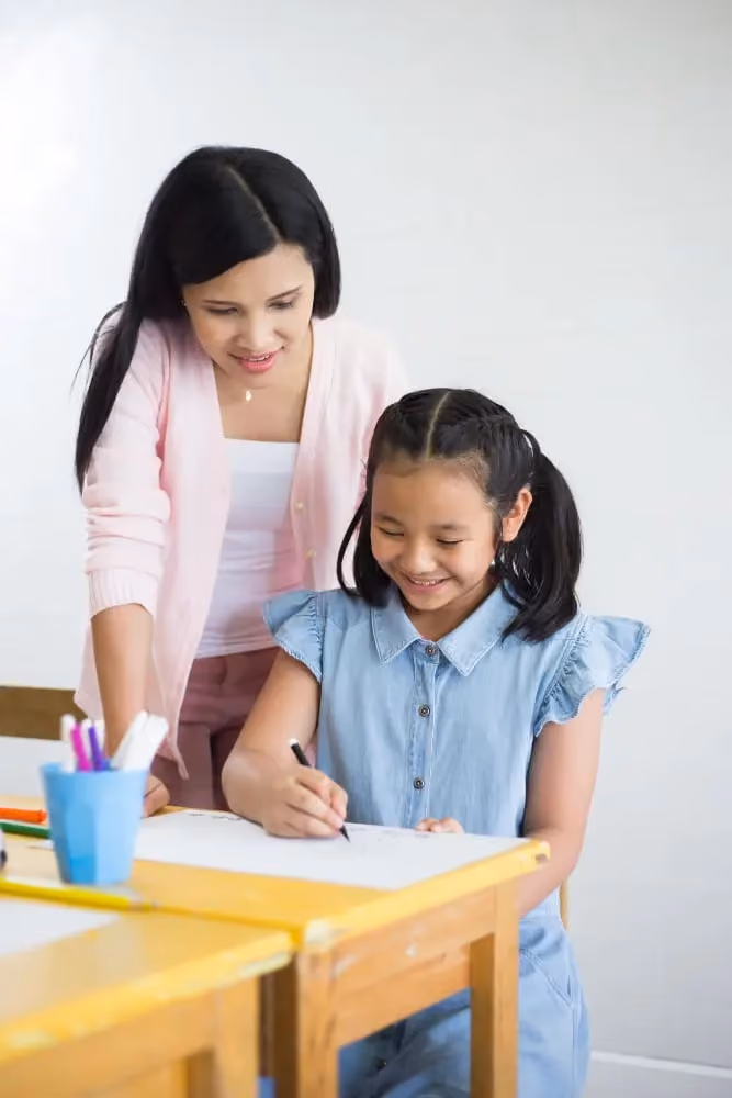 Smiling woman in a pink cardigan guiding a young girl as she writes at her desk in a classroom.