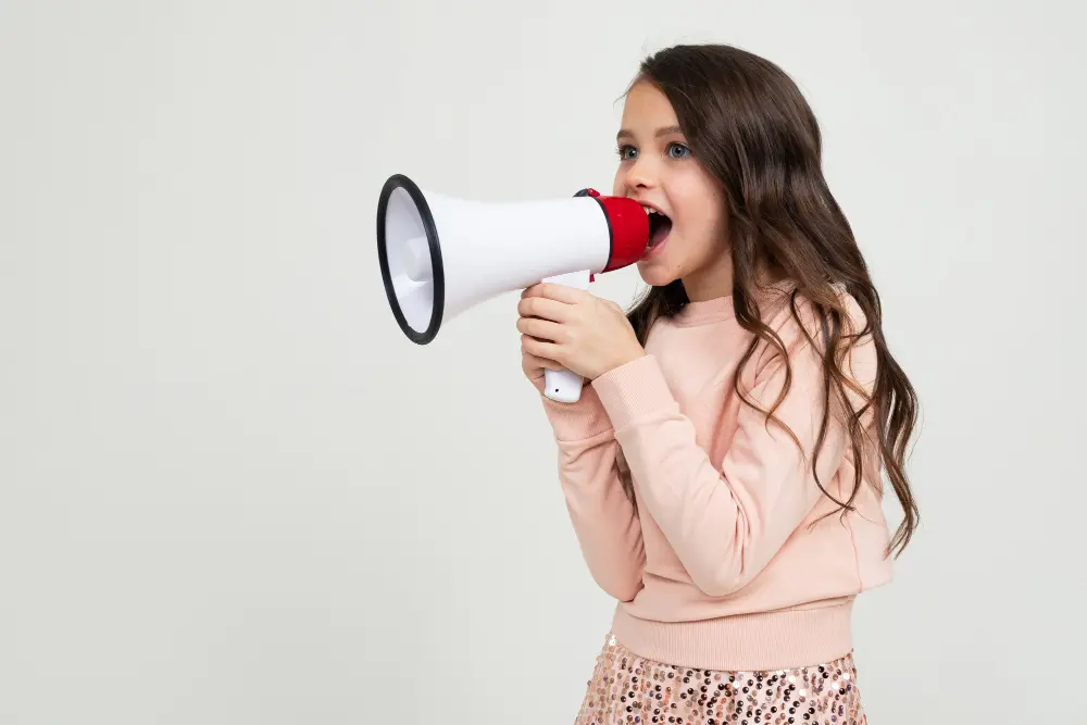 Young girl with long brown hair speaking loudly and confidently into a megaphone on a light background.