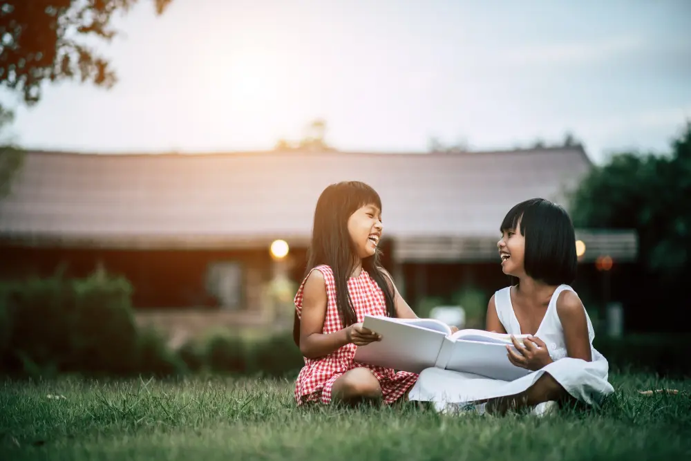 Two young Asian girls sitting on grass laughing and reading books together outdoors in the golden hour.
