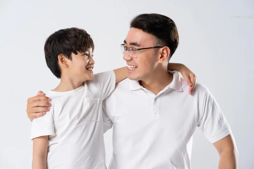 Smiling young Asian man with glasses and a boy with arms around each other, showing mentor-student connection.