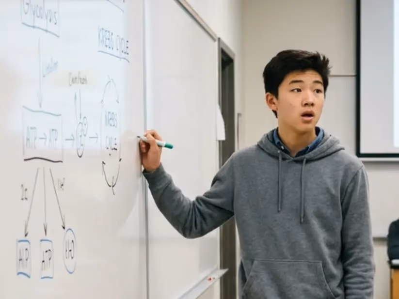 Asian teenage boy in a gray hoodie writing a diagram on a whiteboard while presenting in class.