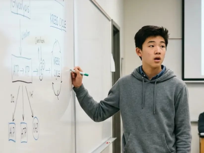 Asian teenage boy in a gray hoodie writing a diagram on a whiteboard while presenting in class.
