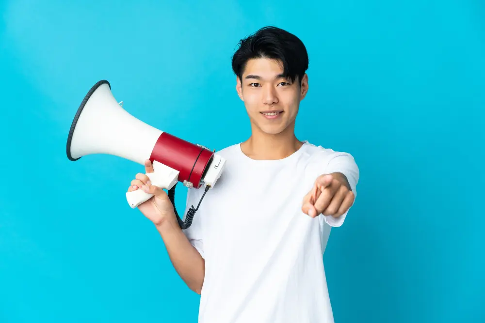 Confident young Asian man holding a megaphone and pointing forward against a blue background.