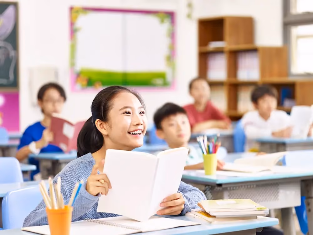 Smiling Asian girl holding an open book and looking up attentively in a bright classroom with other students.