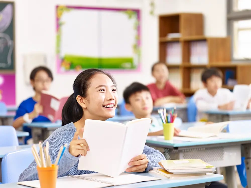 Smiling Asian girl holding an open book and looking up attentively in a bright classroom with other students.