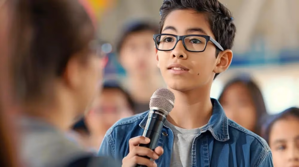Focused teenage boy with glasses holding a microphone and speaking at a school public speaking event.