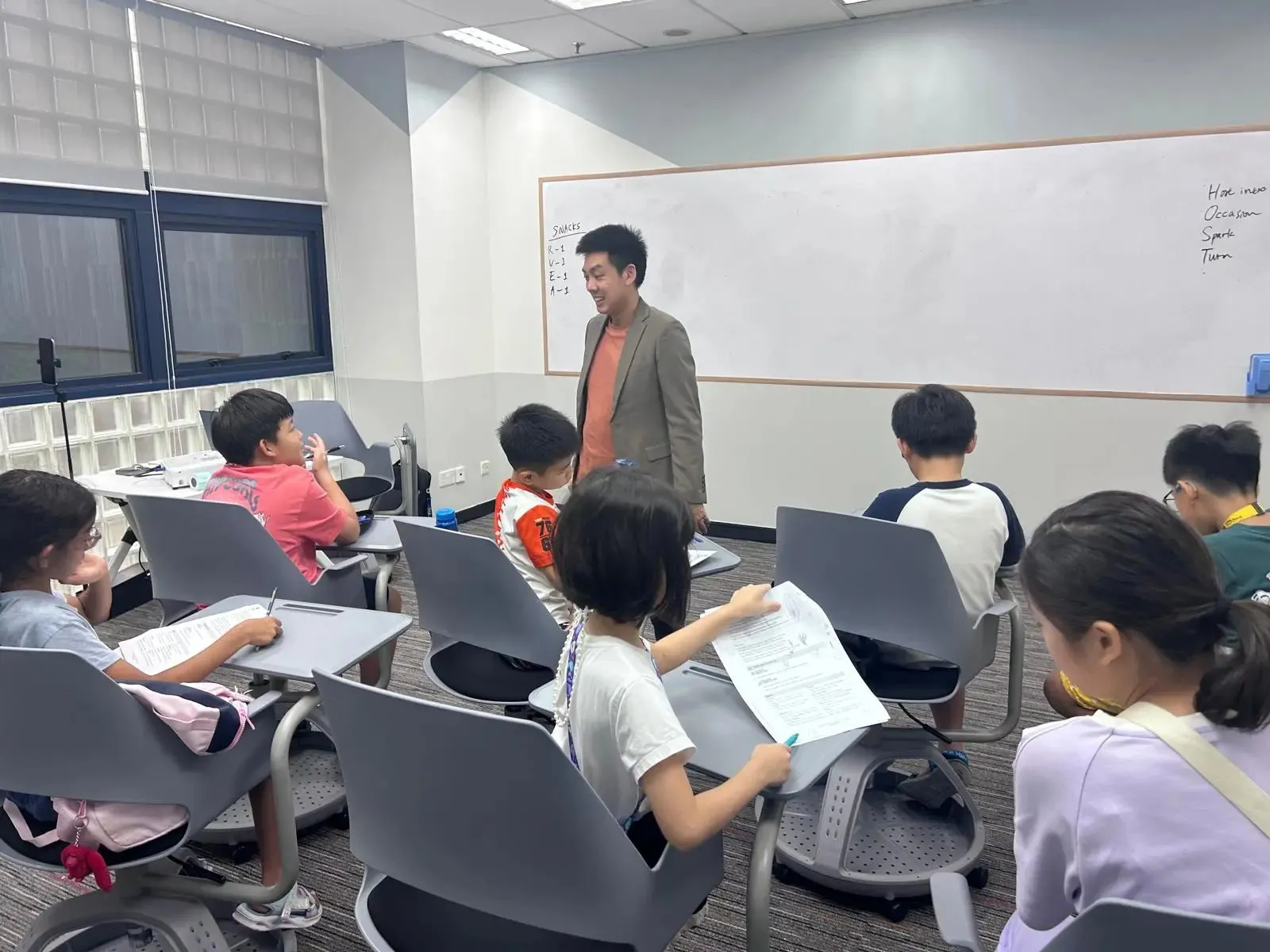 Teacher standing and smiling in front of seated children holding papers in a classroom with a whiteboard.
