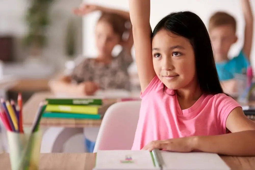 Young Asian girl in a pink t-shirt raising her hand confidently at her desk in a classroom.