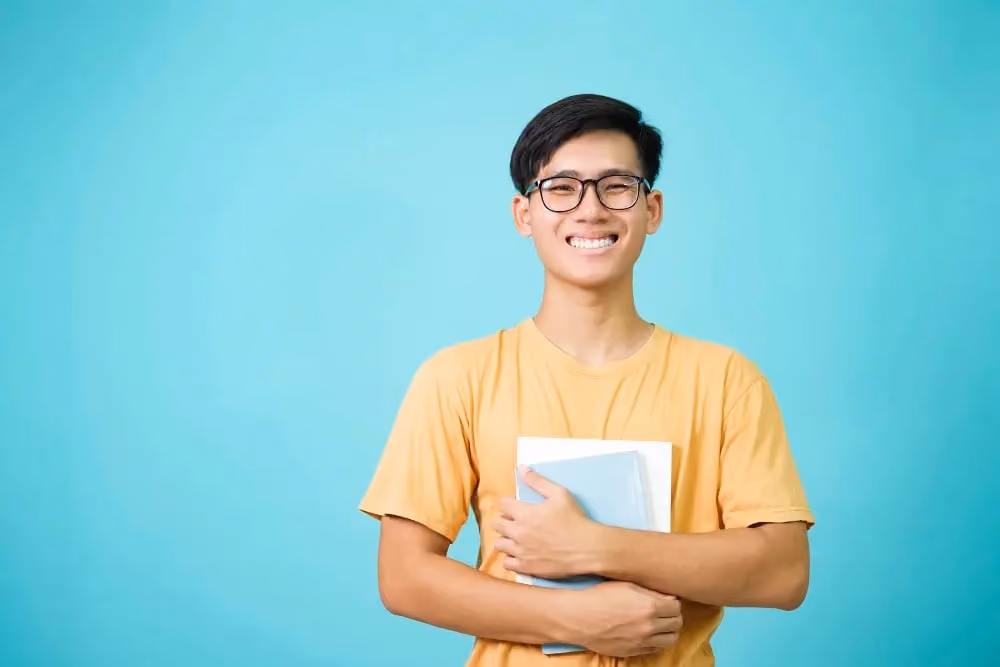 Smiling young man with glasses in yellow shirt holding books against blue background.