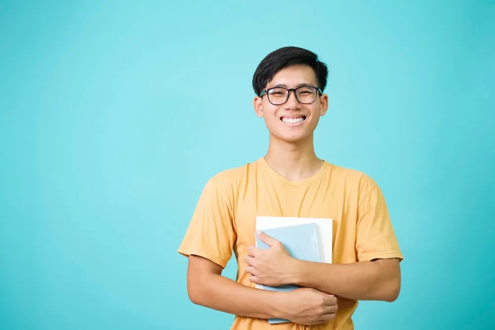 Smiling young man with glasses in yellow shirt holding books against blue background.